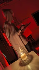 una persona con mantello e cappuccio beige legge un libro di profilo. candela sul tavolo in primo piano. sfondo con luce rossa